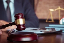 Person im Anzug hält einen Richterhammer, im Hintergrund Justizwaage und Hausmodell.