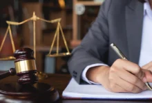 Eine Person im Anzug schreibt auf ein Dokument an einem Schreibtisch mit Richterhammer und Justizwaage.